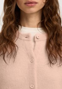 Vrouw met lang, golvend bruin haar die een lichtroze gebreid vest met knopen draagt over een wit topje, afgebeeld van hals tot borst.