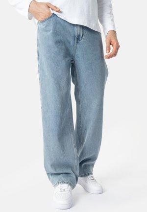 Persona che indossa jeans azzurri a gamba larga, sneakers bianche e una maglia a maniche lunghe bianca con una mano in tasca.