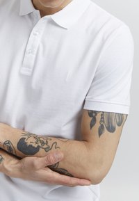 Hvit bomullspolo med treknappslukking og ribbestrikket krage. Inkluderer subtilt merkevarelogo og synlige arm tatoveringer.