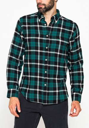 Uomo con camicia a quadri verde, nera e bianca con maniche lunghe e pantaloni neri, in piedi davanti a uno sfondo bianco.