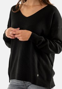 Maglione nero lavorato a maglia con scollo a V, polsini e orlo a costine. Logo a forma di cuore piccolo e distintivo vicino al fondo sul lato sinistro.