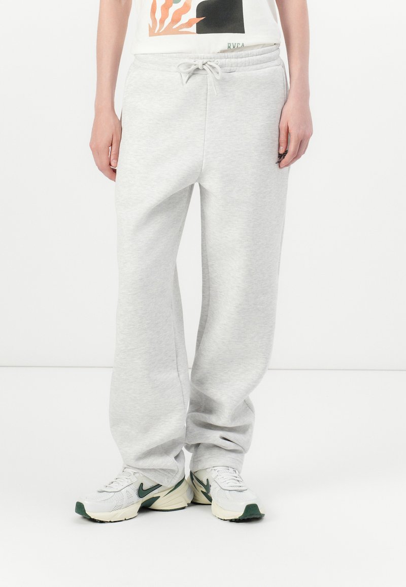 Persoon in lichtgrijze sweatpants met een trekkoord en witte sneakers met donkergroene accenten, staande tegen een witte achtergrond.