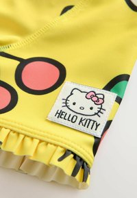 Sárga fürdőruha fekete és rózsaszín kör alakú mintákkal, fehér hímzett "Hello Kitty" címkével és fodros szegéllyel.