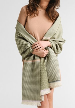 Femme vêtue d'une robe beige enveloppée dans un châle vert à motifs avec des franges, tenant le châle fermé autour de son torse.