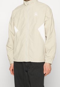 Chaqueta beige con cremallera y cuello alto, con paneles laterales blancos, llevada con pantalones negros, mostrando el torso y los brazos pero sin cabeza.
