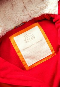 Intérieur d'un manteau en polaire rouge avec une doublure fluffy crème et une étiquette orange. L'étiquette indique "Little Bird" et possède des espaces pour les coordonnées.