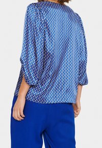 Donna che indossa una blusa blu a fantasia con maniche a sbuffo, abbinata a pantaloni blu, vista da dietro contro uno sfondo chiaro.