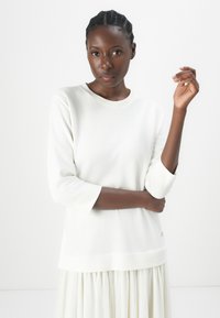 Maglione bianco in cotone con maniche a tre quarti, scollo rotondo e un delicato logo in evidenza all'orlo. Tessuto liscio, vestibilità comoda.