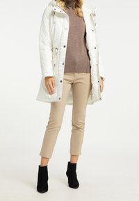 Manteau isolé blanc avec capuche, fermeture à boutons et à glissière, porté sur un pull brun et un pantalon beige, associé à des bottines noires à la cheville.
