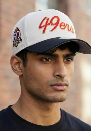 Young man wearing a white and black San Francisco 49ers cap with "60 seasons" patch, looking slightly to the right against a blurred background.