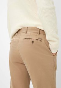 Chinos beige con una texture liscia, dotati di tasche posteriori con chiusura a bottone e un'etichetta con marchio sulla cintura.