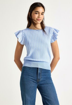 Nuori nainen, joka käyttää vaaleansinistä ribbineulottua toppia, jossa on röyhelöiset lyhyet hihat, ja korkeavyötäröisiä sinisiä farkkuja, seisoo valkoista taustaa vasten.