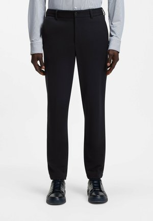Mann trägt schwarze gerade geschnittene Hose, hellgraues langärmeliges Hemd und schwarze Ledersneakers, steht vor einem weißen Hintergrund.
