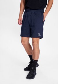 Marineblauwe shorts met een elastische tailleband, zijzakken en een wit logo op de rechteronderkant. Gepaard met zwarte sneakers en sokken.