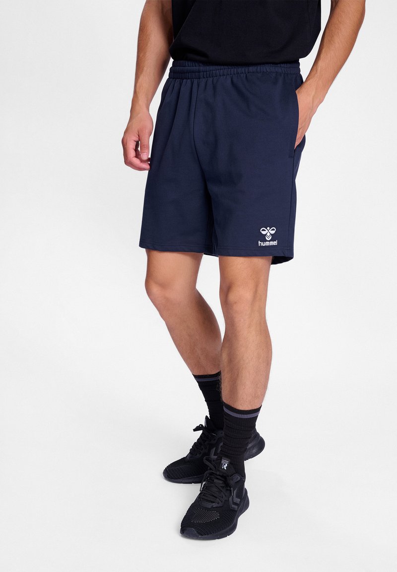 Marineblauwe shorts met een elastische tailleband, zijzakken en een wit logo op de rechteronderkant. Gepaard met zwarte sneakers en sokken.
