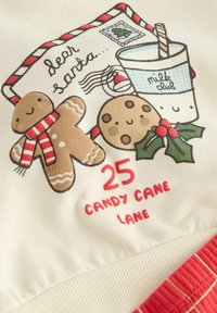 Tkanina w kolorze kremowym z rysunkowym piernikiem, ciastkiem czekoladowym, kubkiem mleka i czerwonym napisem "25 CANDY CANE LANE". Czerwone pasy jako akcenty.