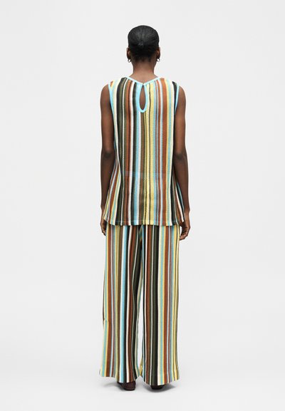 Γυναίκα με σκούρα μαλλιά σε κότσο, που φοράει μπλούζα χωρίς μανίκια και παντελόνι με φαρδιά πόδια με κάθετες ρίγες σε μπλε, κίτρινο, καφέ και άσπρο, στέκεται γυρισμένη προς τα πίσω.