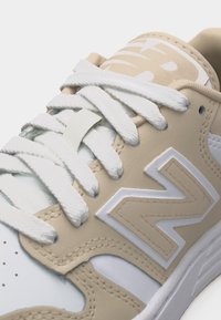 Zapatilla beige y blanca con cordones blancos, un gran logo "N" cosido en el lateral y zona de puntera perforada.