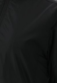 Sort vindjakke med glat, letvægtsstof, lynlås foran og paneldetaljer ved skulderen. Minimalistisk design.