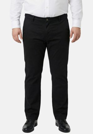 Mand iført sorte slim-fit bukser, sorte pæne sko og en hvid skjorte med knapper, står med armene afslappet ned langs siderne.