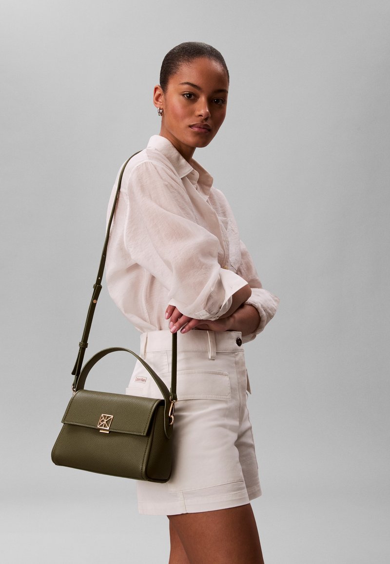 Γυναίκα με λευκό πουκάμισο και σορτς που κρατά μια τσάντα ώμου από δέρμα σε ελιά πράσινη απόχρωση με χρυσό κούμπωμα και ρυθμιζόμενο λουρί.