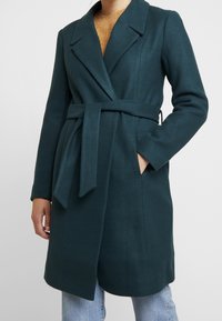 Kvinde iført en mørkegrøn uldfrakke med bælte over en brun sweater og lyseblå jeans, med den ene hånd i frakkens lomme.