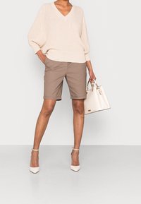 Suéter beige de punto oversize con un escote profundo en V, combinado con pantalones cortos de corte recto en marrón claro y un bolso blanco. Los zapatos son pálidos y con punta.