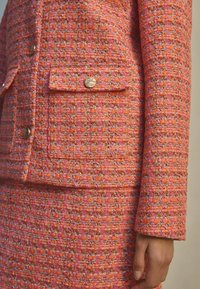 Gestructureerde blazer van roze en oranje tweed met een zak. Voorzien van accenten met knopen in goudkleur. Getailleerde pasvorm met zichtbare stikdetails.