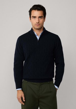 Pull en tricot côtelé bleu marine avec une fermeture éclair quart, des poignets et un ourlet côtelés. Porté par-dessus une chemise col bleu clair, associé à un pantalon vert olive.