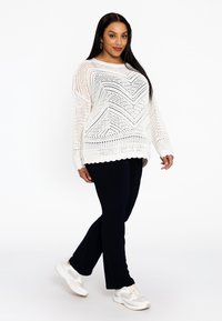 Weißer Strickpullover mit geometrischem Muster und Wellenkante, kombiniert mit marineblauen Hosen und weißen Sneakers. Einfaches und strukturiertes Design.