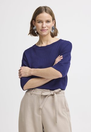 Maglione a coste di colore blu navy con scollatura ampia e maniche corte, abbinato a pantaloni beige a vita alta con cintura legata.