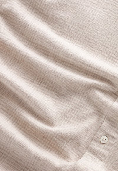 Lys beige tekstureret stof med et subtilt ternet mønster, prydet med en hvid knap i kanten og blødt, draperet udseende.