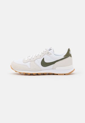 Baskets Nike blanches et beiges avec un swoosh et un renfort au talon vert olive, semelle en caoutchouc naturel, et lacets blancs, présentées de profil sur fond blanc.
