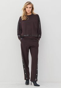 Moteris vilkinti tamsiai rudą treniruočių kostiumą su juodomis blizgiomis juostelėmis ir mygtukais ant kelnių, derinant su juodais aukštakulniais bateliais iki kulkšnies, stovi prieš baltą sieną.