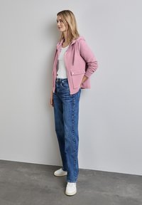 Pink hooded jacket with two front pockets, paired with light blue straight-leg jeans and white sneakers. Model stands against a neutral background.