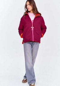 Kobieta w bordowej pikowanej kurtce z różowym zamkiem błyskawicznym i jasnoszarymi dżinsami, stojąca z dłońmi w kieszeniach i skrzyżowanymi nogami.