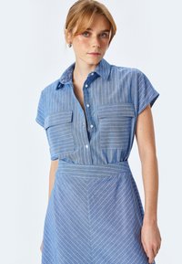 Abito camicia blu con maniche corte e collo a punta. Presenta strisce verticali bianche, due tasche sul petto e una vita arricciata.