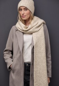Beige gebreide sjaal met ribbelstructuur, om de hals gedrapeerd, gecombineerd met een lichtgrijze jas, een crème shirt en een bijpassende beige gebreide muts.