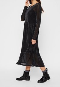 Vestido negro de manga larga, hecho de una tela transparente y texturizada con un acabado brillante. Corte en A con dobladillo acampanado, combinado con botas negras de tobillo.