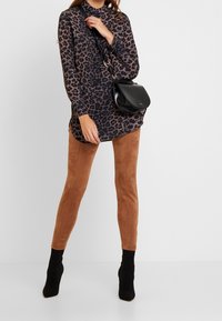 Femme portant un chemisier à imprimé léopard, des leggings en suède marron, des bottines chaussettes noires, et un sac bandoulière en cuir noir.