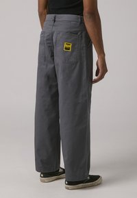 Gray, wide-leg pants with a smooth texture, featuring a back pocket and a yellow branded label. Paired with black sneakers.