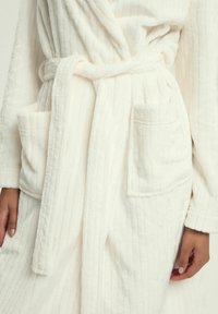 Biały pluszowy szlafrok z pionowym prążkowaniem, z dwoma przednimi kieszeniami i pasującym paskiem, wykonany z miękkiego, teksturowanego materiału.