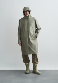 Zielony wodoodporny płaszcz z zamkiem błyskawicznym, regulowanym kapturem i bocznymi kieszeniami. W zestawie z teksturowanymi, oliwkowozielonymi kaloszami z wzorem.