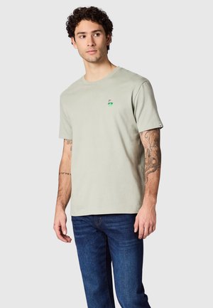 Man met krullend haar die een lichtgrijs t-shirt draagt met een klein groen golfvlag-embleem en blauwe spijkerbroek, staand tegen een effen achtergrond.