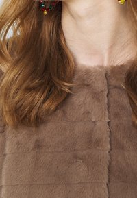 Bruin nep bont kledingstuk met een gestructureerde afwerking, met een vierkant ontwerp en een ronde halslijn. Het haar is golvend en omlijst het gezicht.