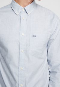 Košile na knoflíky ve světle modré látce s kostkovaným vzorem, s náprsní kapsou a vyšitým logem na levé straně.