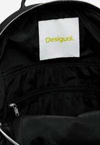 Interieur van een zwarte nylon rugzak met een wit label waarop "Desigual." in het geel staat. Inclusief een ritsvak en een gladde stoftextuur.