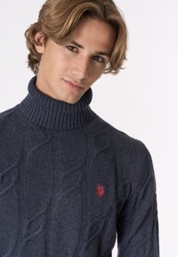 Giovane uomo con capelli castano chiaro che indossa un maglione a collo alto blu scuro con lavorazione a trecce e un logo ricamato rosso.