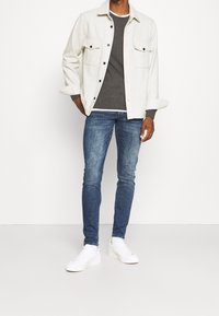 Chemise bouton-up gris clair avec deux poches avant, pull gris foncé, jean bleu et baskets blanches. Textures lisses et coupe décontractée.