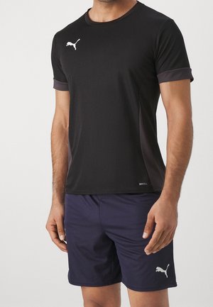 Mand iført sort Puma DryCELL kortærmet atletisk skjorte og marineblå Puma shorts, stående med armene afslappet.